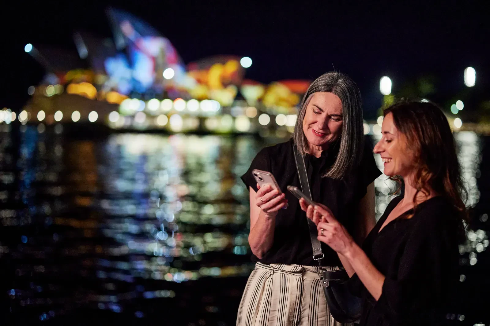 Two individuals standing by the water at night, illuminated by the lights of the Sydney Opera House in the background. They are both engaged with their smartphones, reflecting a blend of digital interaction and cultural engagement in a vibrant, nocturnal setting.