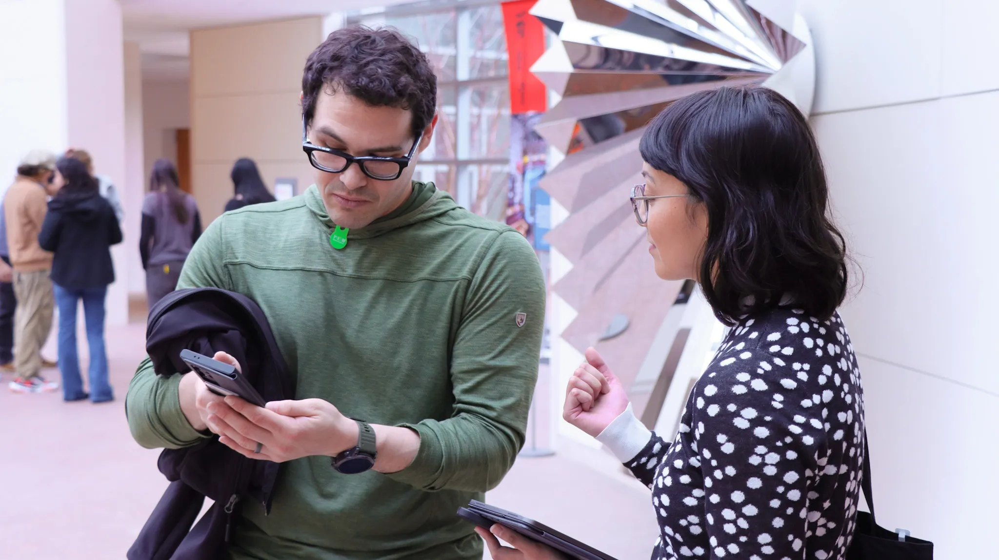 A researcher conducts on-site user testing with a visitor inside a cultural venue. The visitor holds a phone and a jacket while reviewing something on the screen, as the researcher gestures and holds a tablet. Other visitors and a reflective sculpture are visible in the background, indicating an active public space.