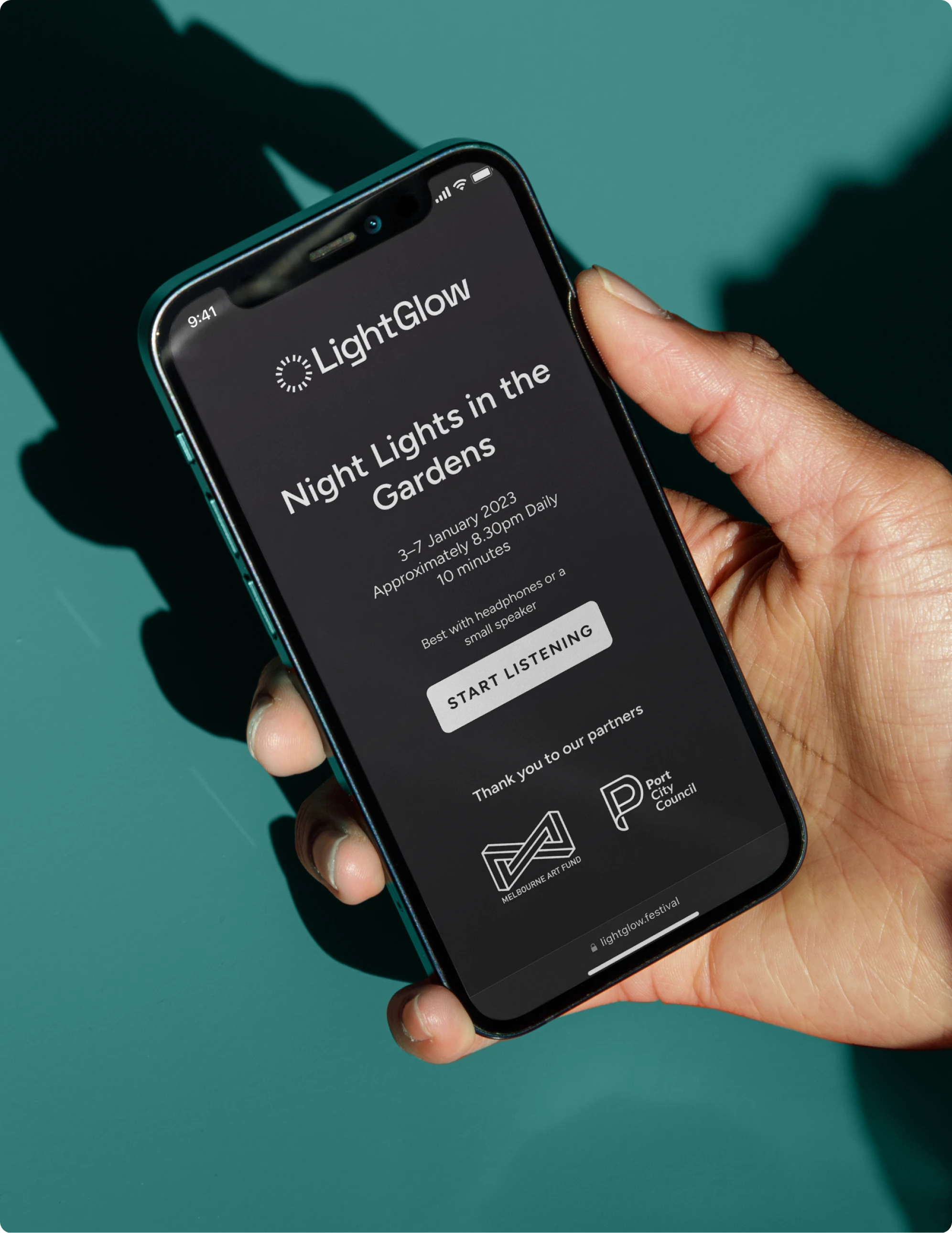 A person holds a smartphone displaying the Event Sync interface for “Night Lights in the Gardens,” an outdoor event running from January 3–7. The screen shows event details, listening instructions, a start button, and partner logos for Melbourne Art Fund and Port City Council, emphasizing digital audio guidance for park visitors.