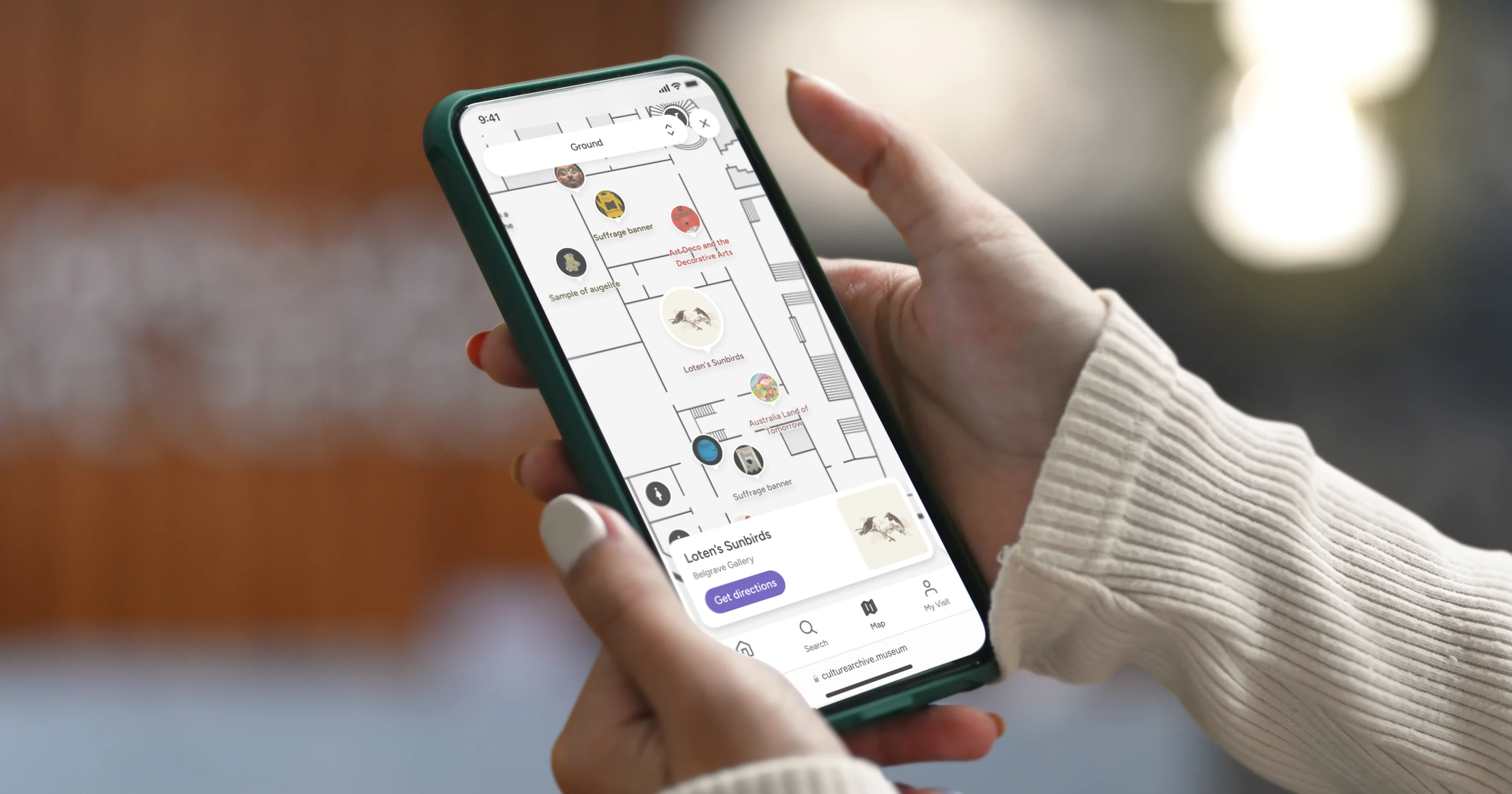 A person holding a smartphone displaying a digital map of an art venue with highlighted rooms and artwork icons, offering directions to specific pieces within the exhibition space.