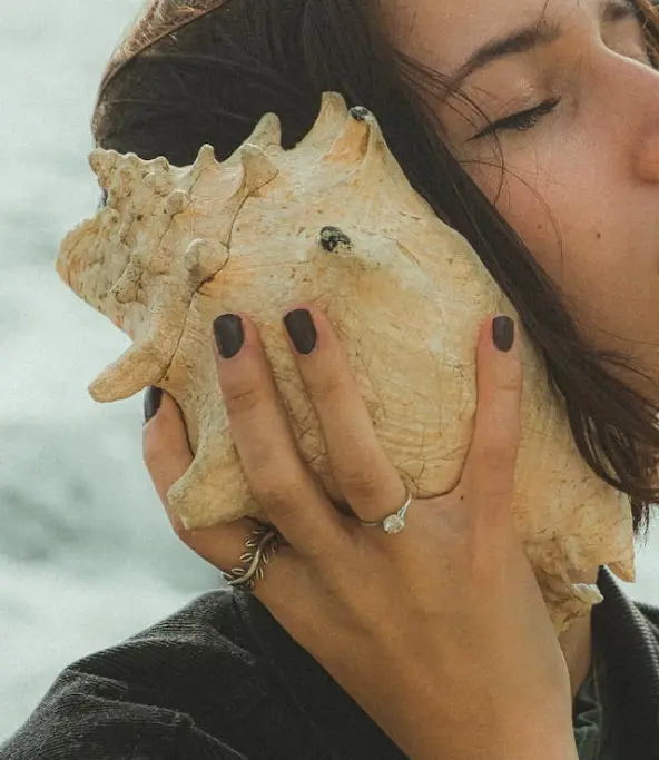 Femme tenant un coquillage près de l’oreille, moment de calme et d’écoute des sons de la mer.
