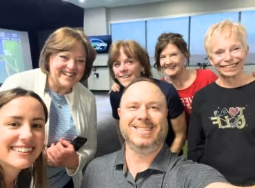 Group selfie of three women and one man smiling indoors with a TV screen and windows in the background.