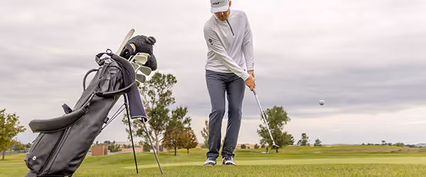A golf bag resting on the green while a golfer chips a shot toward the hole, focusing on precision.
