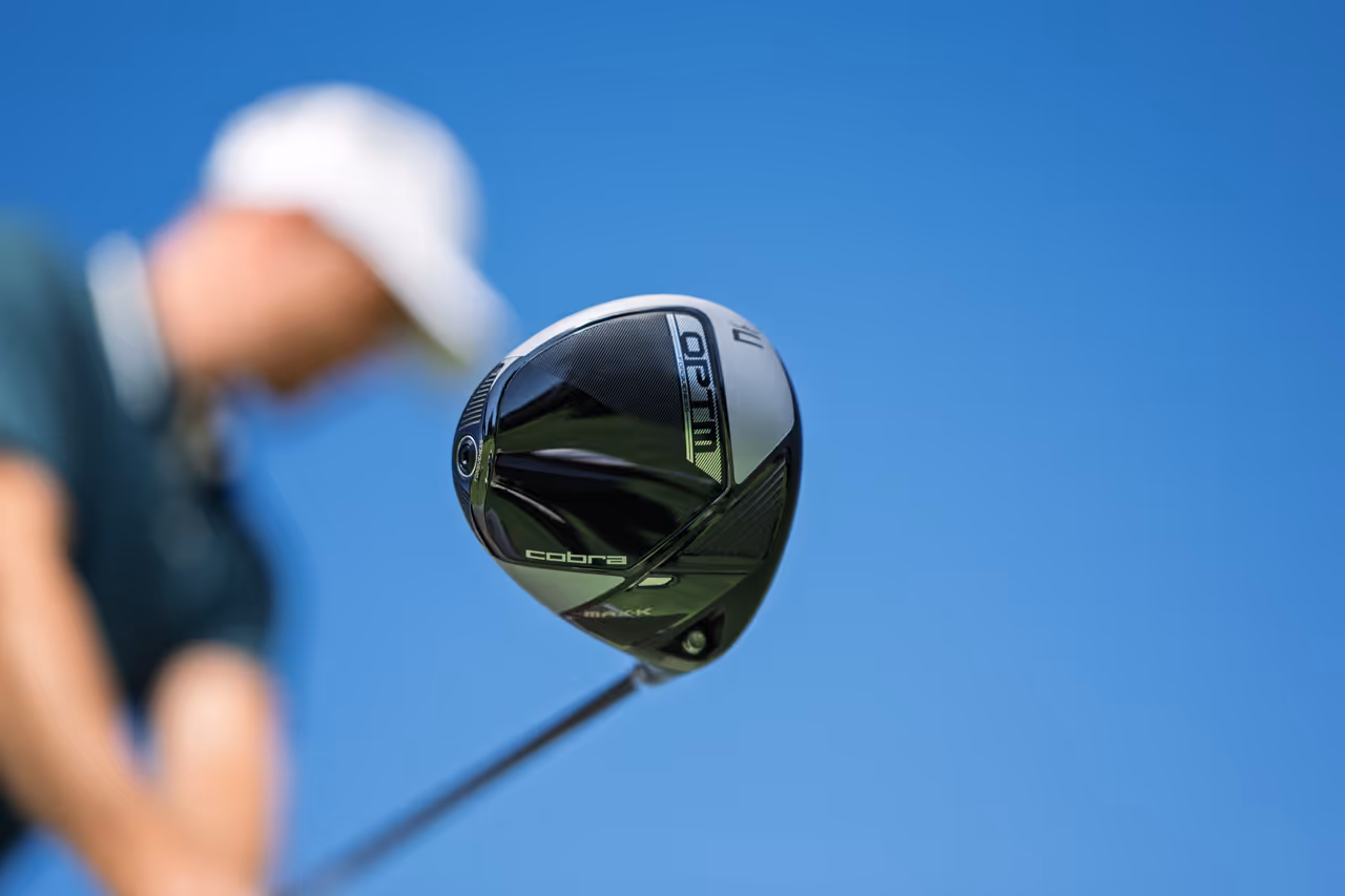 Close-up of a Cobra golf driver with a golfer in a white cap blurred in the background against a clear blue sky.