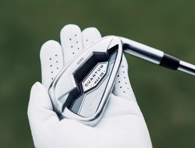 A gloved hand holding a Callaway Quantum Max OS golf iron against a green background.