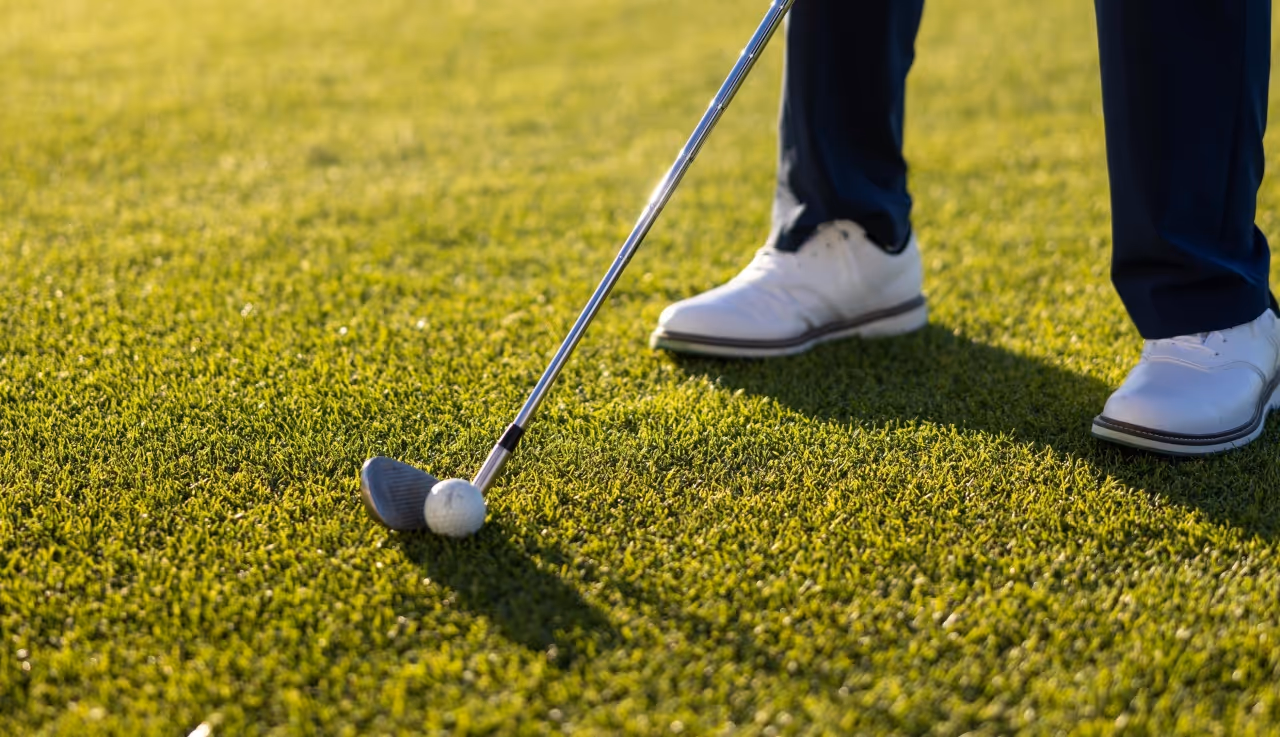Close-up of a golfer's white shoes and golf club ready to hit a golf ball on green grass.