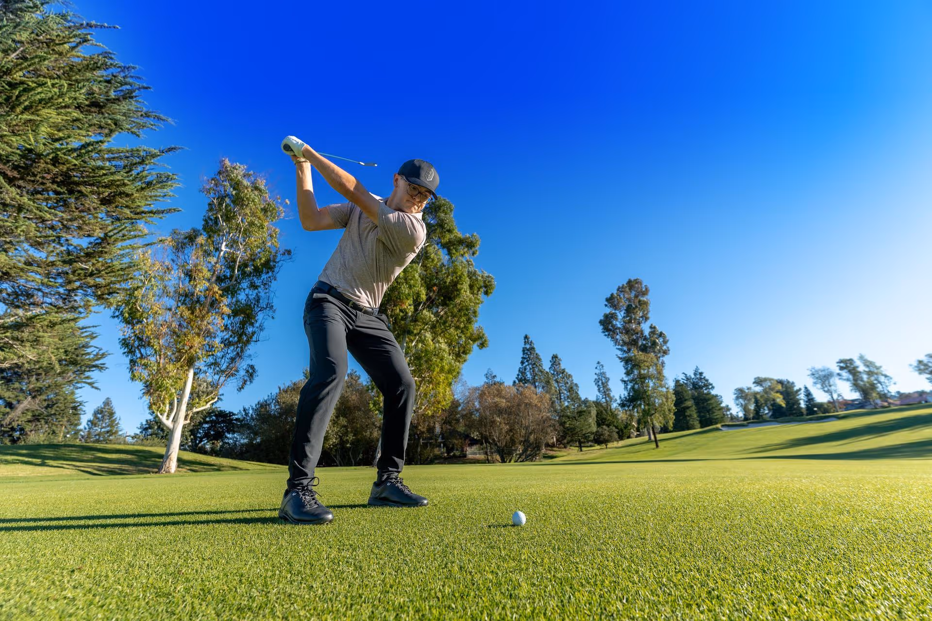Man in glasses swinging golf club on a sunny golf course with green grass and trees.