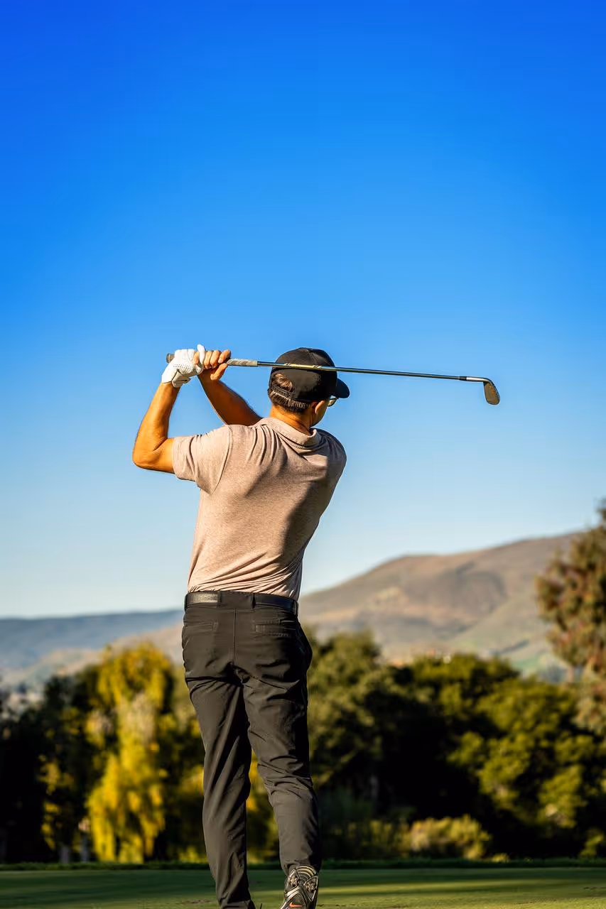Golfer in a light brown shirt and black cap swinging a golf club on a sunny golf course with trees and hills in the background.