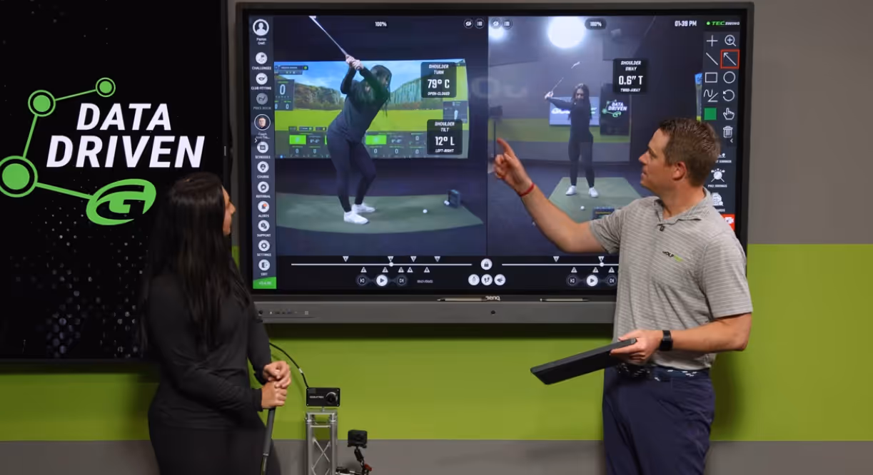 A golf instructor and student analyzing golf swing data on a large screen with digital metrics and video playback.