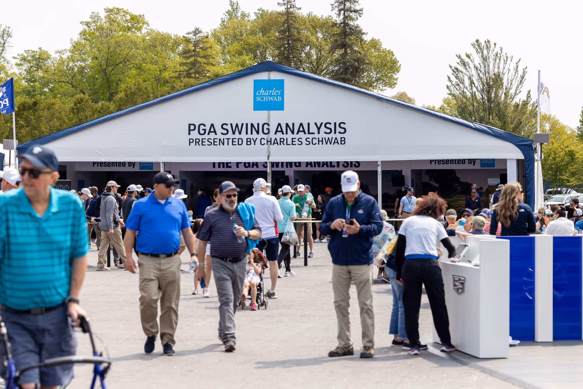 Outdoor event tent with signage 'PGA Swing Analysis presented by Charles Schwab' and people walking and interacting around it.