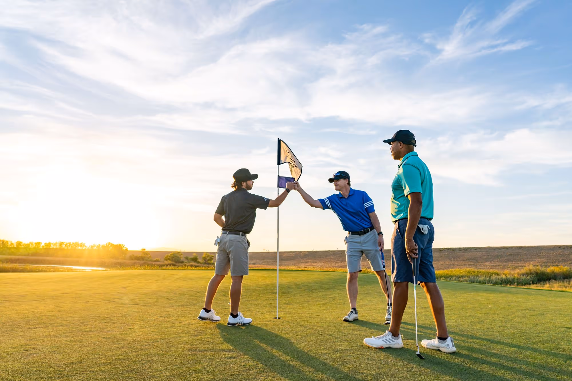 Three golfers on a green at sunset, two fist bumping near the hole flag and one standing with a putter.