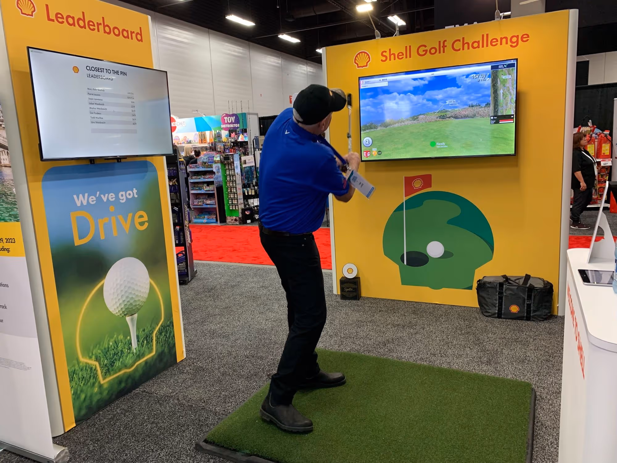Man swinging a golf club at an indoor Shell Golf Challenge with screens showing virtual golf and a leaderboard.
