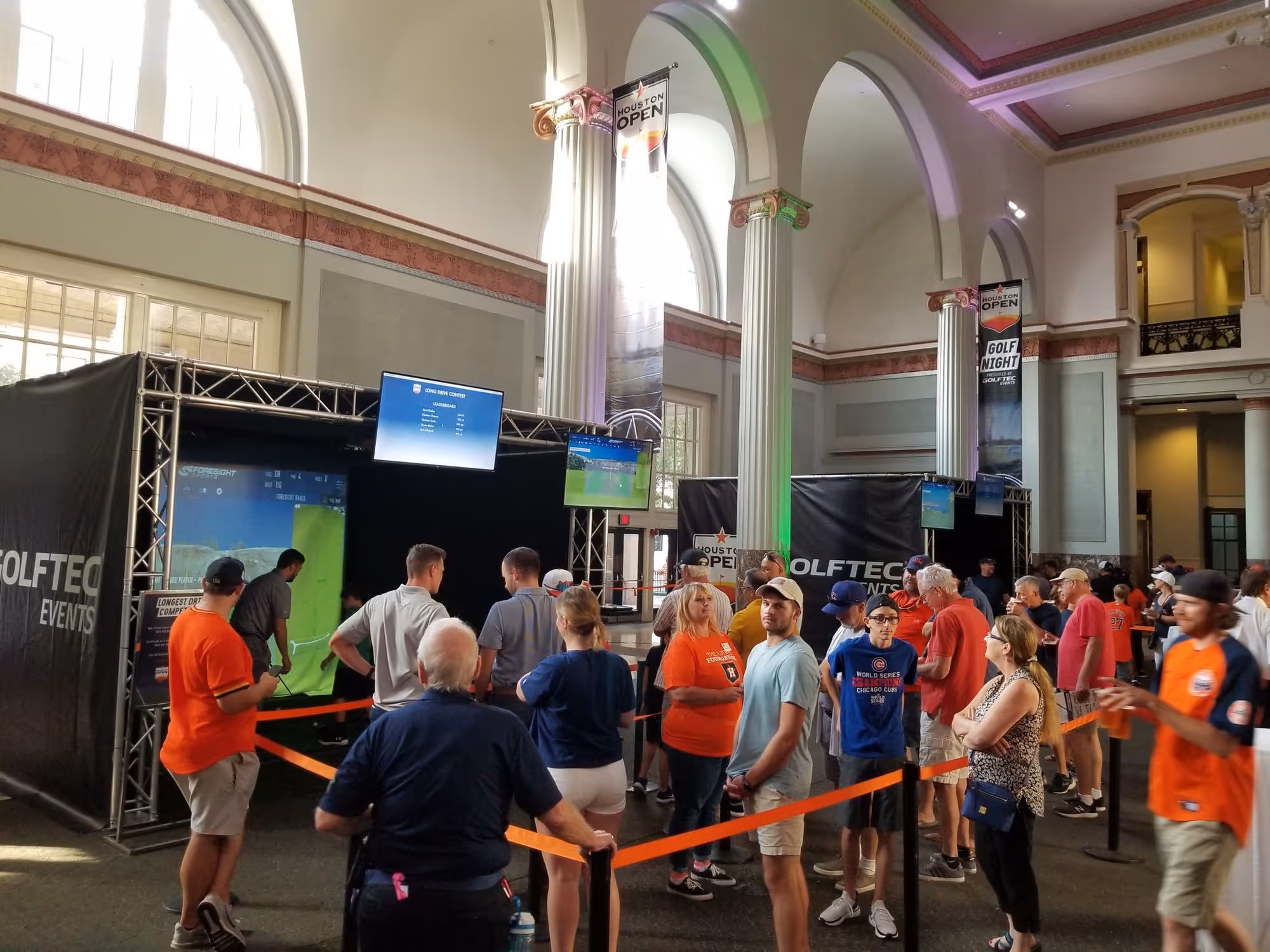 People lined up inside a building with high arches and columns, waiting to use GolfTec simulation stations with screens displaying golf data and the Houston Open banners.