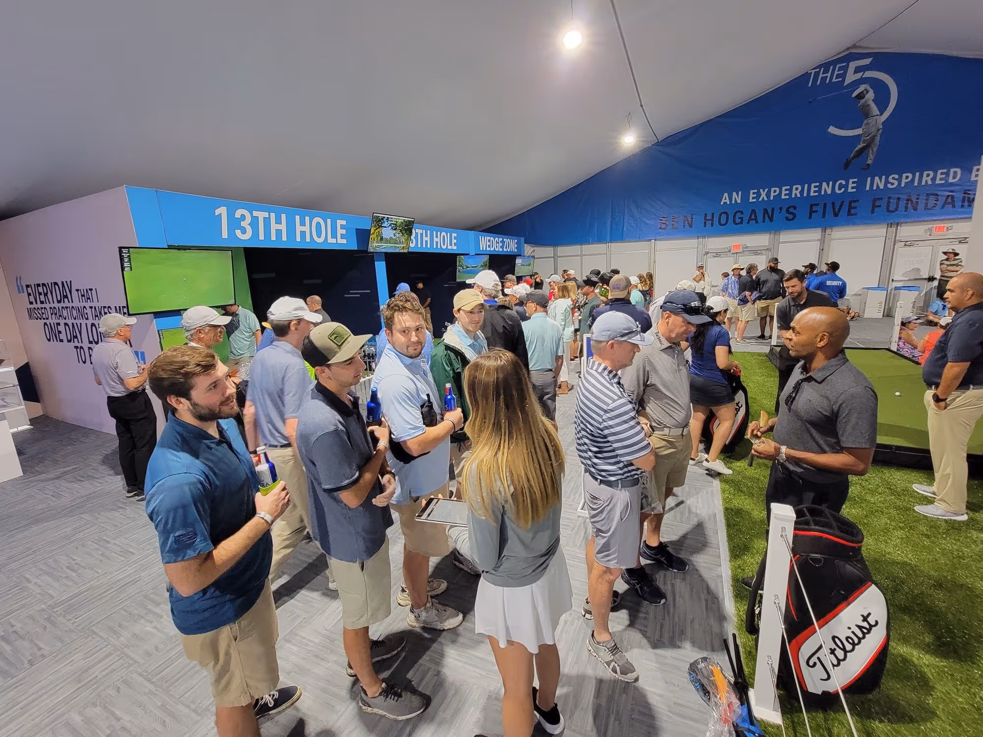 Indoor golf simulation event with people lined up and engaging near stations labeled 13th Hole and Wedge Zone, featuring golf bags and putting green.