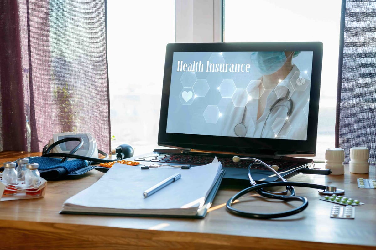 Medical Workspace with Laptop Displaying Health Insurance Information, Surrounded by Stethoscope, Blood Pressure Monitor, Medication Bottles, and Document on Wooden Desk Near Window with Curtains