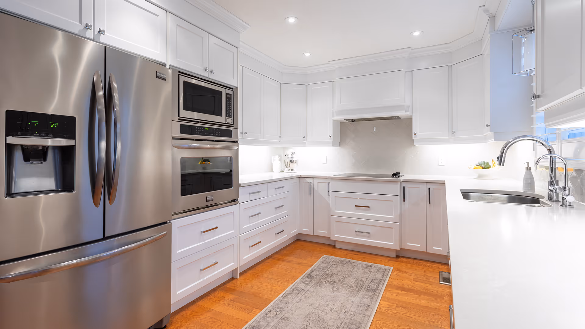 Modern white kitchen with stainless steel refrigerator, built-in microwave and oven, white countertops, and wooden flooring with a light gray rug.