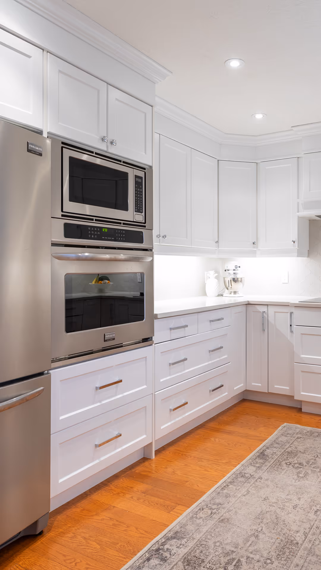 Modern kitchen corner with white cabinets, stainless steel double oven and refrigerator, hardwood floor, and a gray patterned rug.