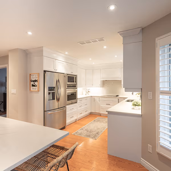 Modern white kitchen with stainless steel refrigerator, built-in oven and microwave, hardwood floor, and a woven chair at a white countertop.