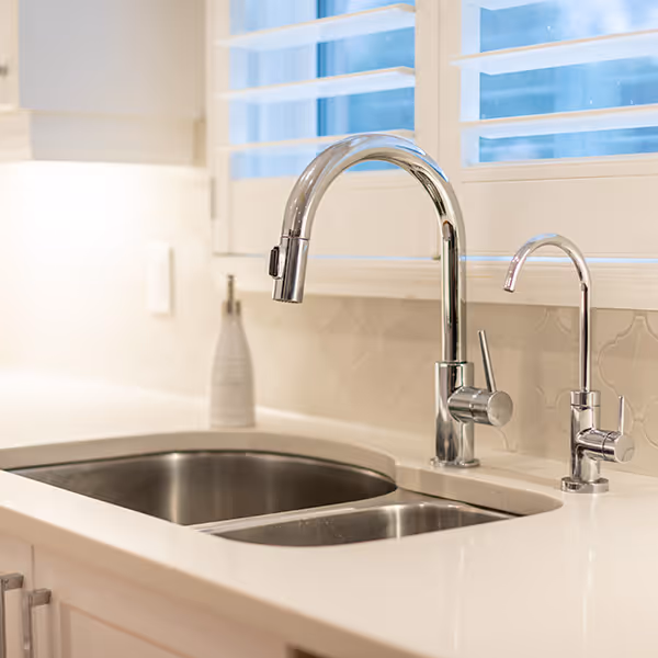 Modern kitchen sink with dual stainless steel basins and a tall curved chrome faucet under white shuttered window.