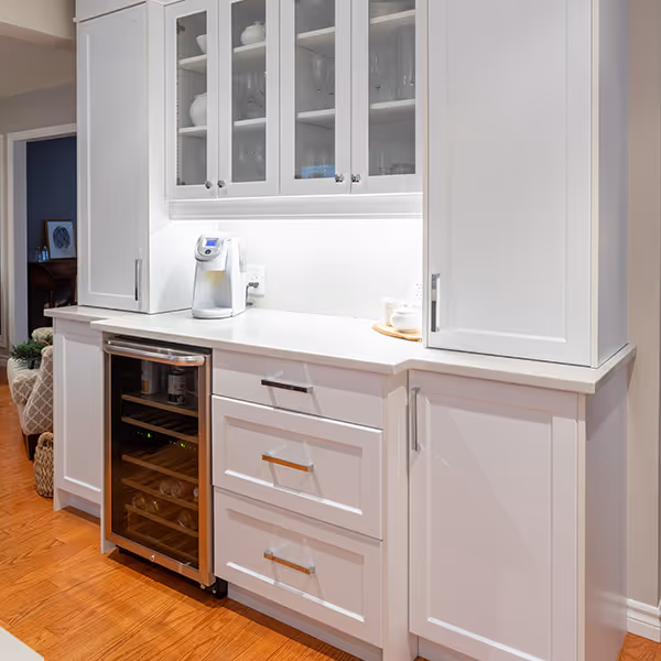 White kitchen cabinetry with glass upper cabinet doors, a counter with a coffee maker, and a wine cooler beneath the counter on a wooden floor.