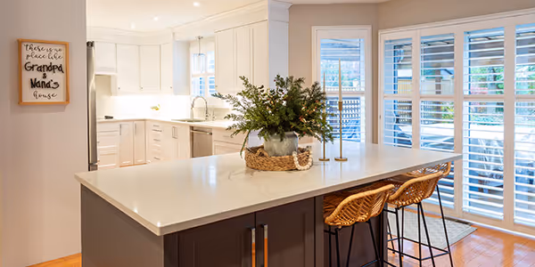 Modern kitchen with white cabinetry, large island with dark base, two wicker bar stools, and large windows with white shutters.