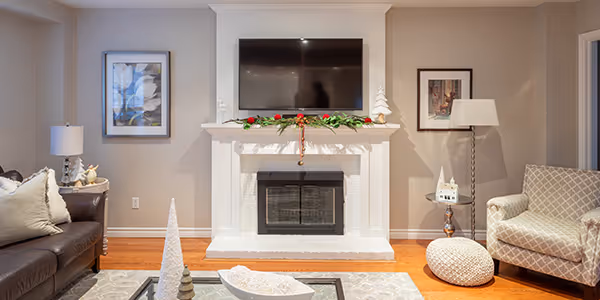 Cozy living room with a white fireplace decorated with greenery and red berries, a mounted TV above, a brown leather sofa on the left, and a beige patterned armchair on the right.