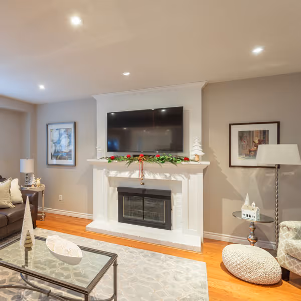 Cozy living room with a white fireplace decorated with greenery and red roses, a mounted flat-screen TV above it, a leather sofa, framed artwork on the walls, glass coffee table, and soft lighting.