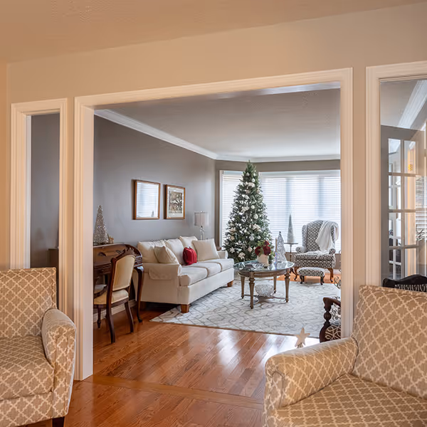 Cozy living room with cream sofa, patterned armchairs, wooden floor, and a decorated Christmas tree near a large window.