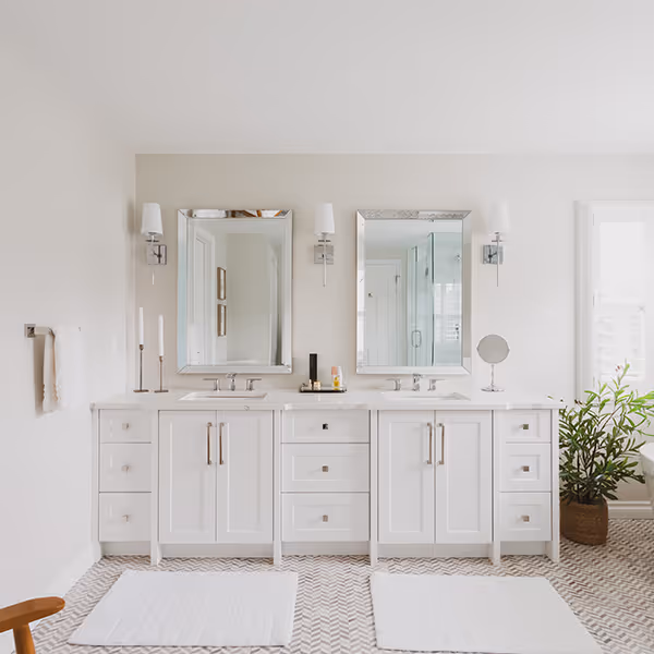 A bright modern bathroom with double vanity sinks, two rectangular mirrors, wall sconces, white cabinetry, and patterned tile floor.