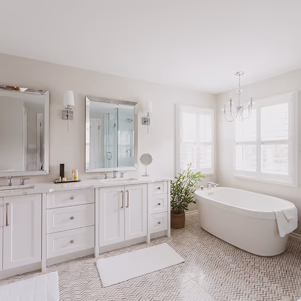 Bright modern bathroom with white dual sinks, large mirrors, a freestanding bathtub, a potted plant, and patterned floor tiles.