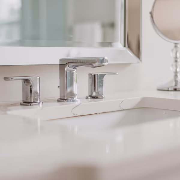 Close-up of modern chrome bathroom faucet and handles on a white sink countertop with a mirror and a round makeup mirror in the background.