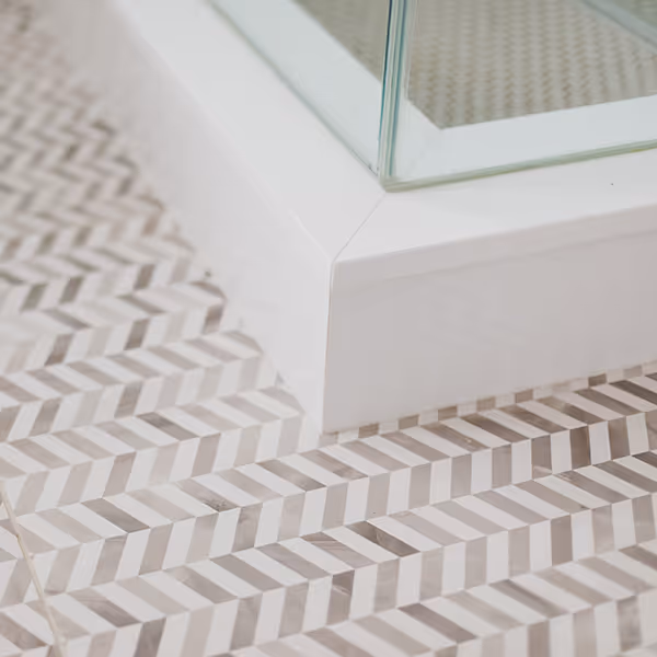 Close-up of a corner where a white baseboard meets glass and patterned floor tiles with a geometric chevron design.