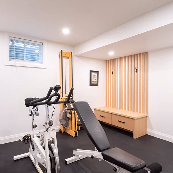 Basement home gym with stationary bike, rowing machine, adjustable bench, and wooden wall-mounted storage bench with hooks.