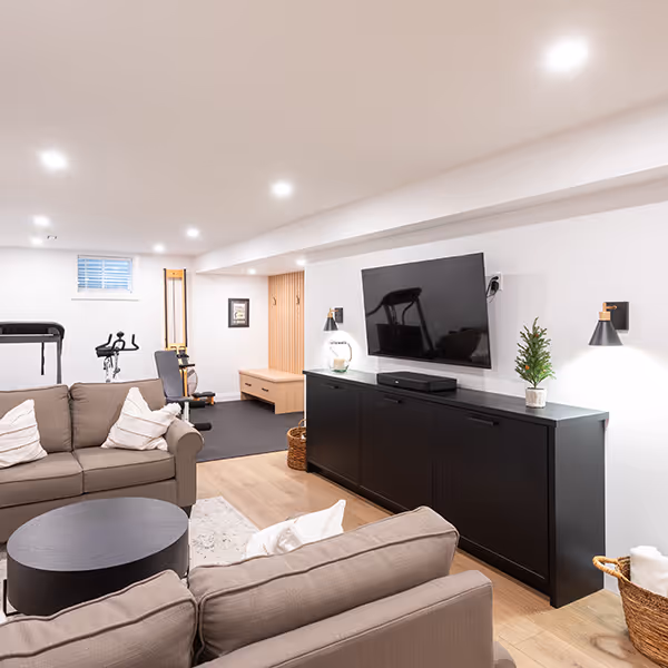 Modern basement living area with beige sofas, black media console with TV, and home gym equipment in the background.