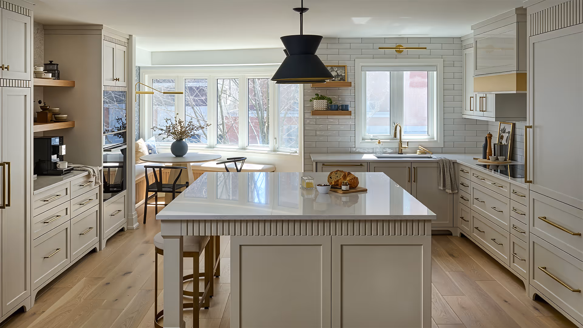 Bright modern kitchen with large white island, gold fixtures, wooden floor, bay window seating, and black pendant light.