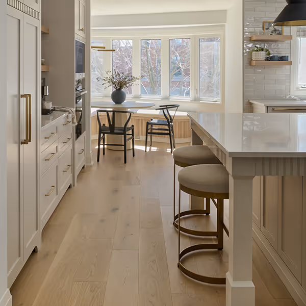 Bright modern kitchen with light wood flooring, white cabinetry, a marble countertop island with two round stools, and a dining nook by large windows with black chairs and a vase of flowers.