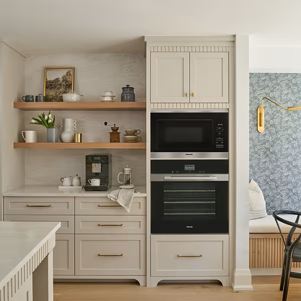 Modern kitchen cabinetry with built-in microwave and oven, open wooden shelves with mugs, coffee grinder, and decor items.