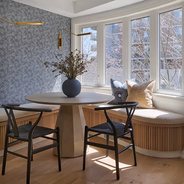 Cozy breakfast nook with round table, two black chairs, built-in bench with cushions, and vase with leafy branches under large windows.