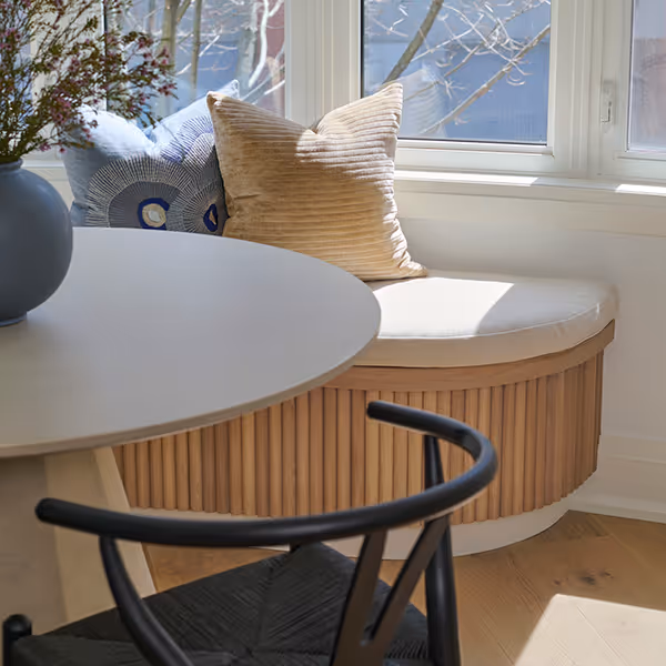 Cozy breakfast nook with curved wooden bench, white round table, black chair, and pillows by a sunlit window.