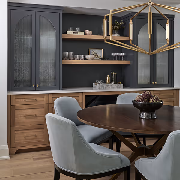 Modern dining area with a round wooden table, four light blue upholstered chairs, grey and wooden cabinetry with glass doors, open wooden shelves, and a gold geometric chandelier.