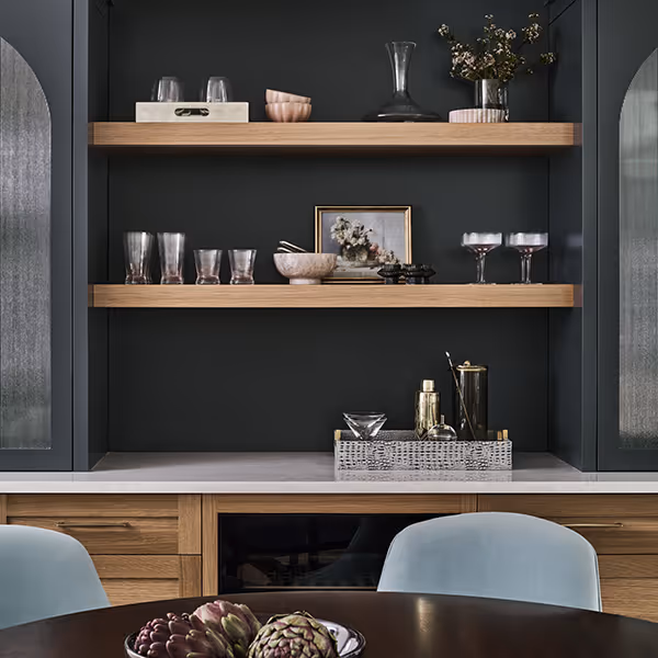 Modern kitchen shelving with wooden floating shelves displaying glassware, a small framed photo, bowls, a decanter, and flowers, above a countertop and wooden cabinets, with a dining table and chairs in the foreground.