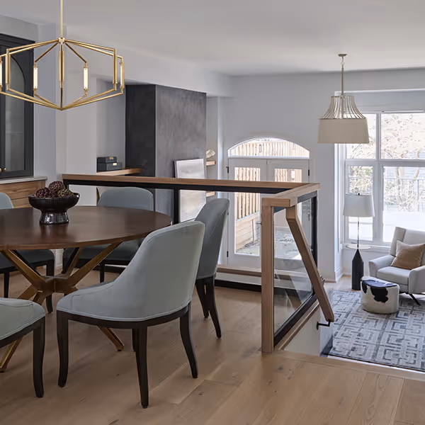 Modern dining area with a round wooden table, blue upholstered chairs, and geometric gold chandelier, adjacent to a sunlit living room with armchair and large windows.