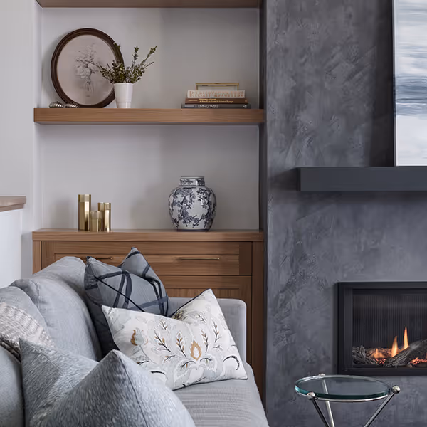 Modern living room corner with a gray sofa adorned with patterned pillows, a wooden shelf with decorative items, and a lit fireplace with a textured gray wall.