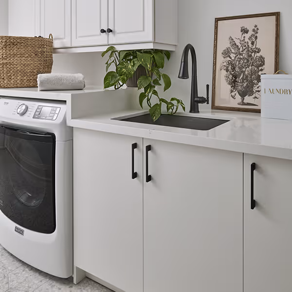 Modern laundry room with white cabinets, black sink faucet, washing machine, a green potted plant, and a framed botanical print.