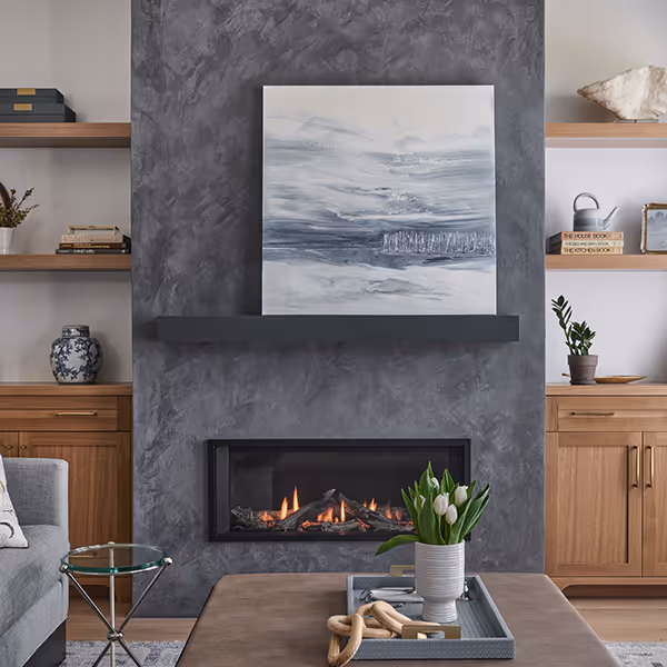 Modern living room with a gray textured fireplace, abstract artwork above, wooden shelves and cabinets, and a coffee table with a vase of white tulips.