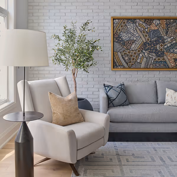Modern living room with a beige armchair, a gray sofa with patterned pillows, a floor lamp, a potted plant, and a framed abstract artwork on a white brick wall.