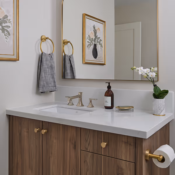 Modern bathroom vanity with white countertop, wood cabinets, gold hardware, a large mirror, gray hand towel, soap dispenser, and white orchid in a marble vase.