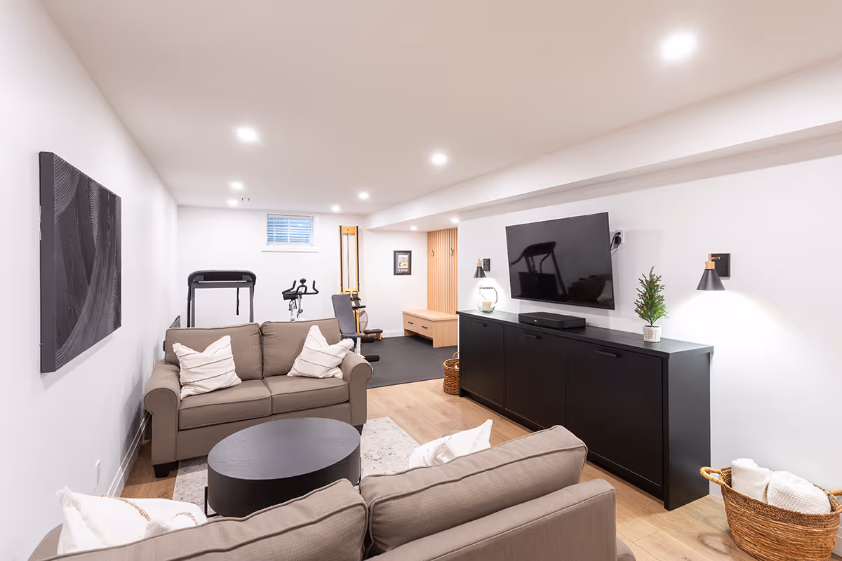 Modern basement living area with beige sofas, black round coffee table, wall-mounted TV, and exercise equipment in the background.
