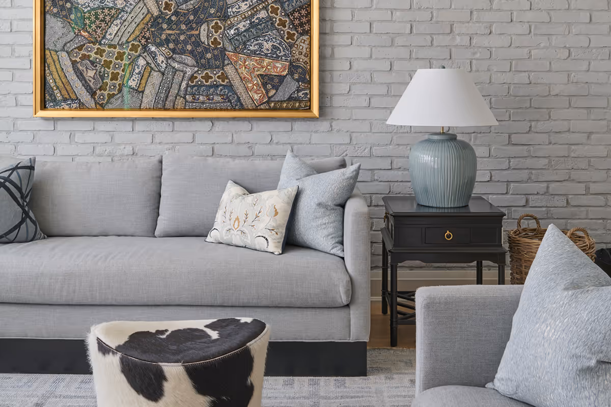 Modern living room with gray sofas, a black and white cowhide ottoman, black side table with a blue ceramic lamp, and a framed abstract artwork on a gray brick wall.