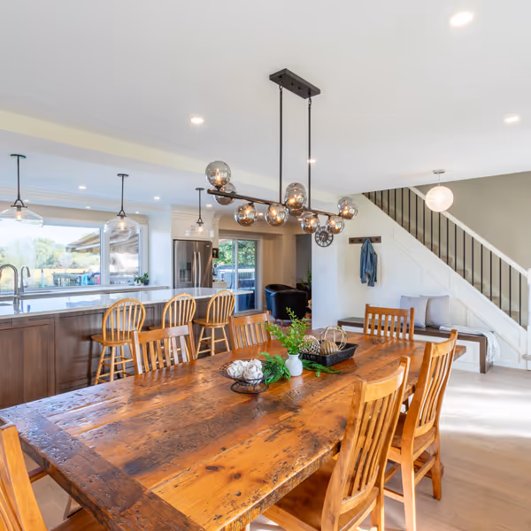 Cozy breakfast nook with round table, two black chairs, built-in bench with cushions, and vase with leafy branches under large windows.